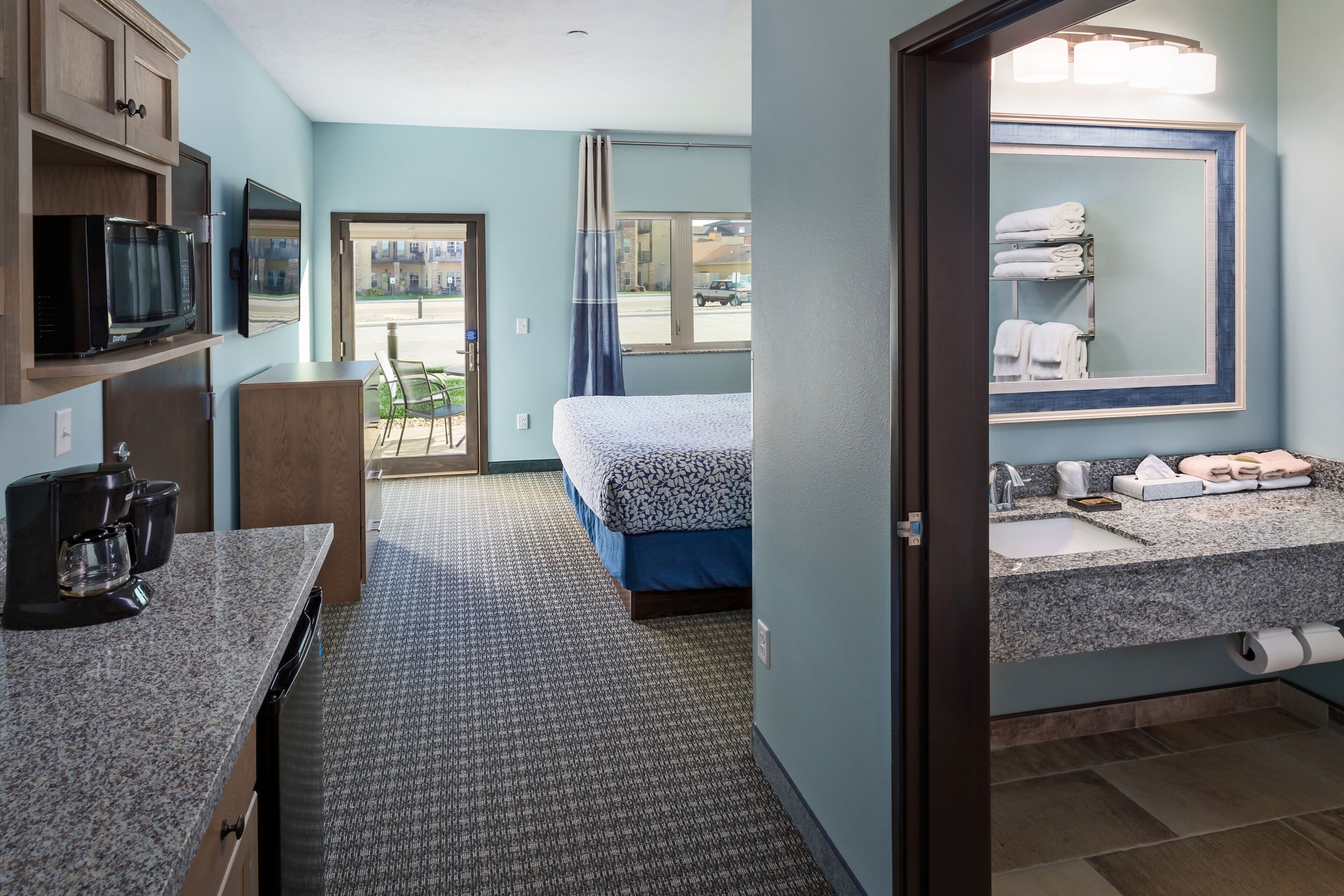 A hotel room with a bed, kitchenette, bathroom, and a view of the outside.