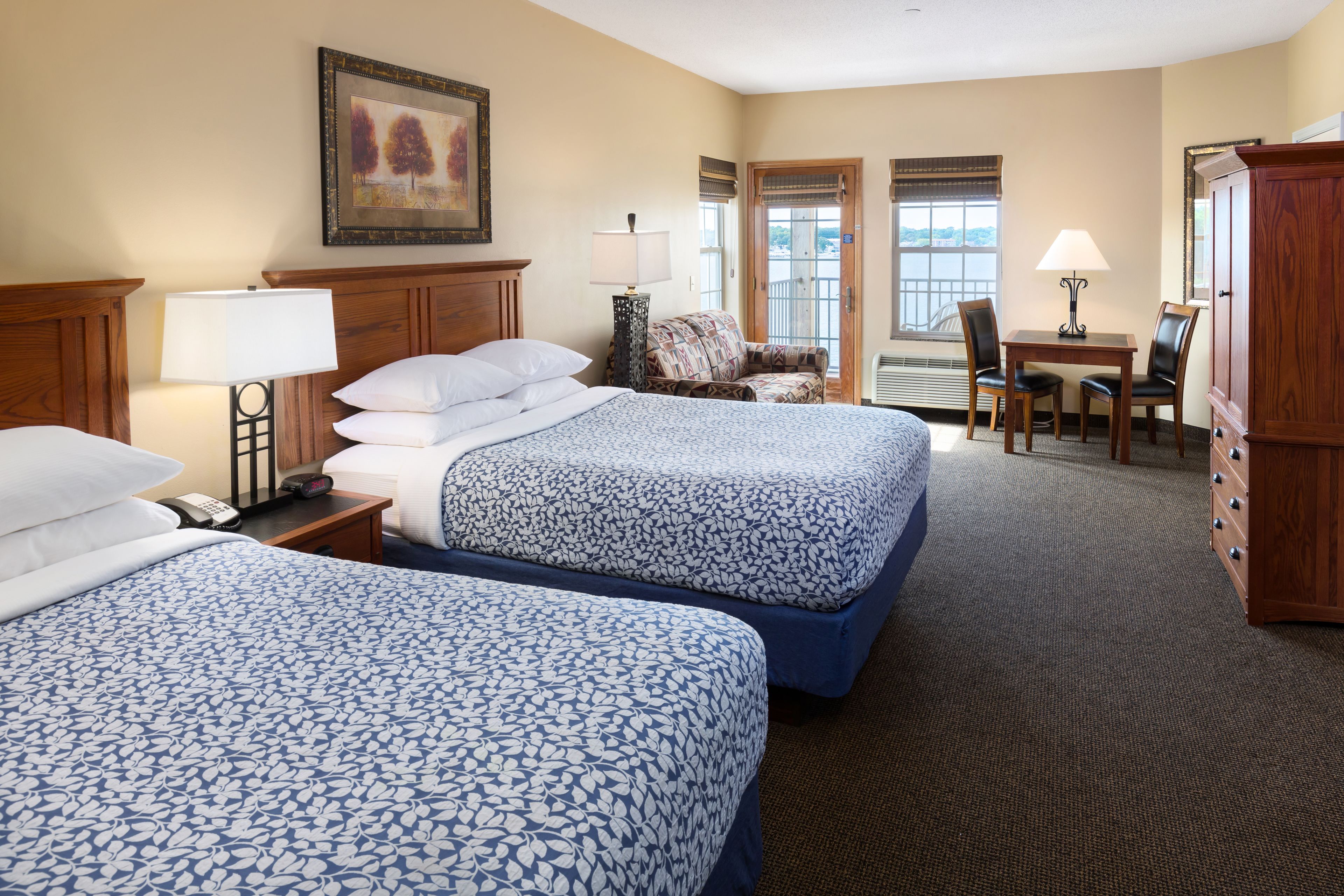 A hotel room with two beds, a desk, a chair, a wardrobe, and a painting on the wall.
