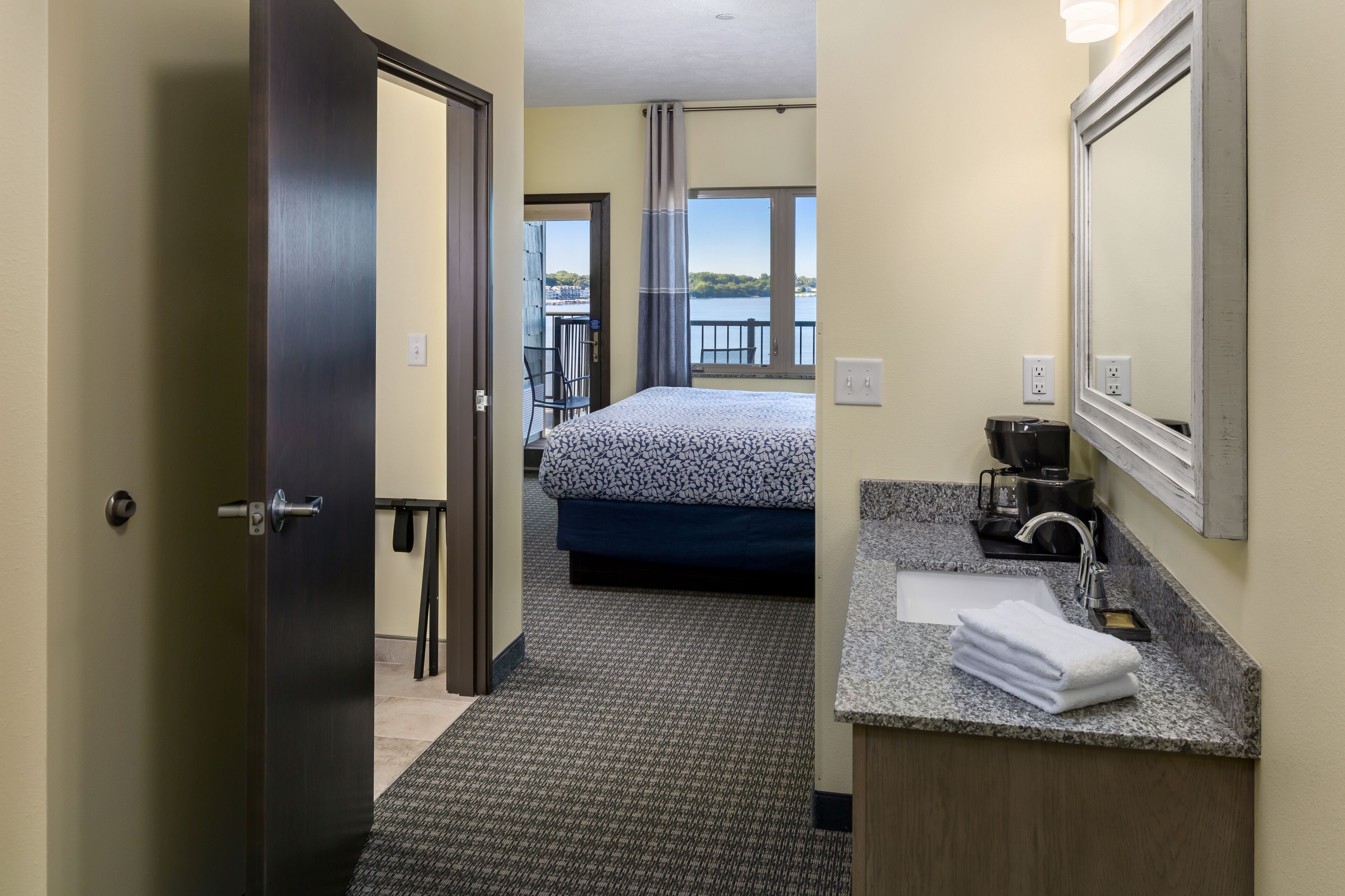 A hotel room with a bed, a bathroom with a sink and mirror, and a balcony with a view of the water.