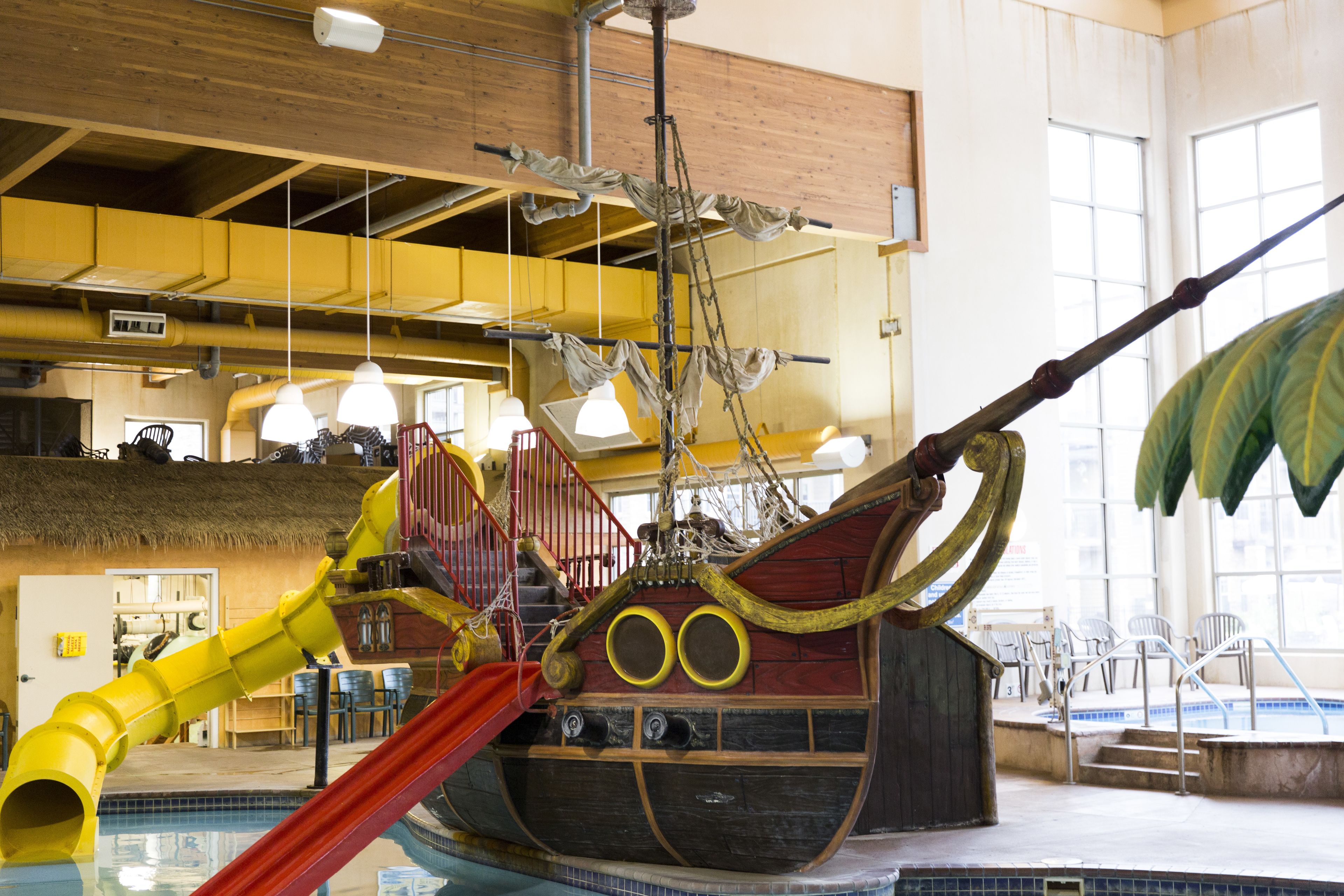 An indoor pool area with a pirate ship play structure, slides, and a thatched-roof seating area.