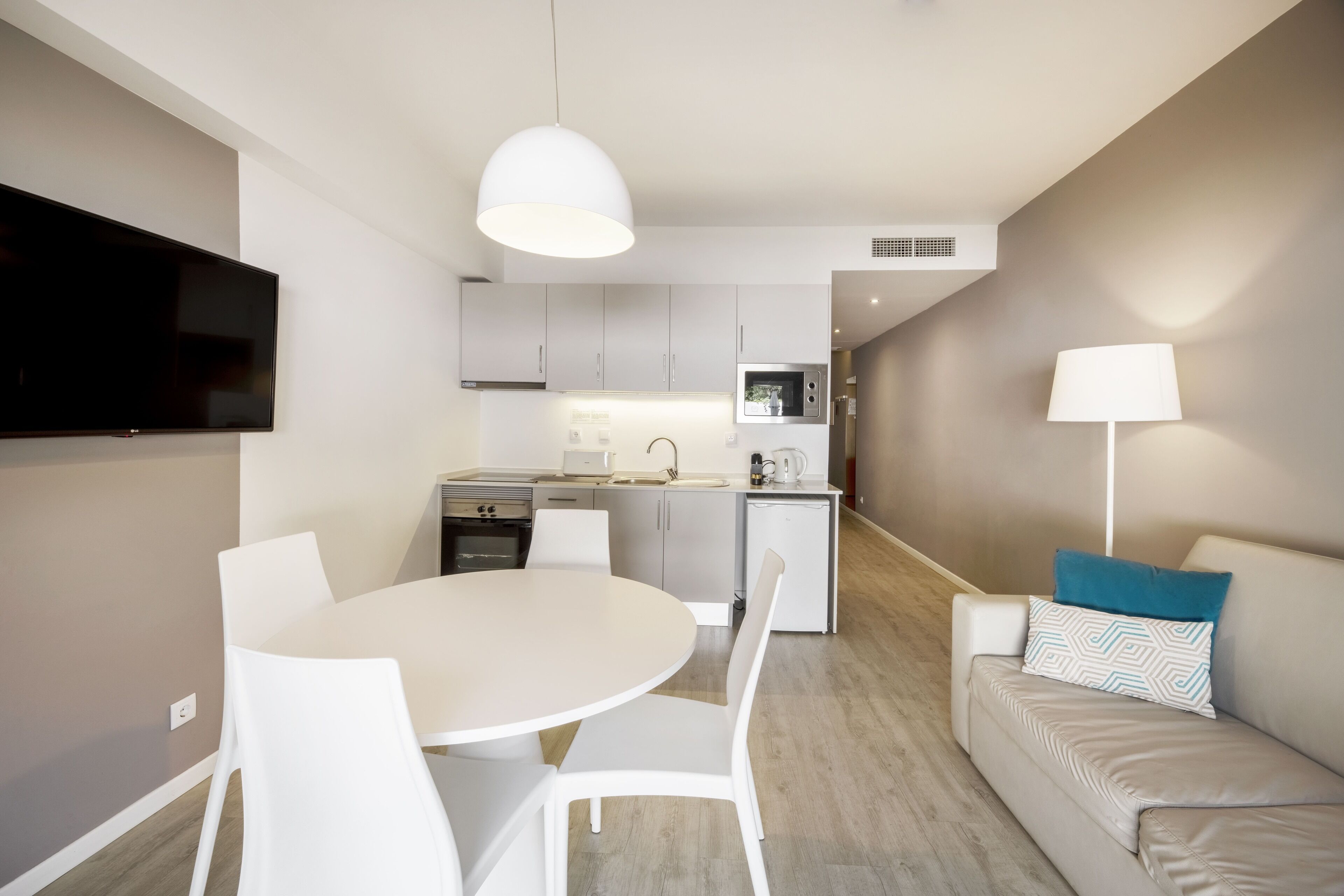 A modern kitchen with white cabinets, a built-in oven, and a microwave. There is a dining area with a white table and chairs, and a sofa with a lamp and pillows.