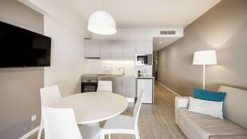 A modern kitchen with white cabinets, a built-in oven, and a microwave. There is a dining area with a white table and chairs, and a sofa with a lamp and pillows.