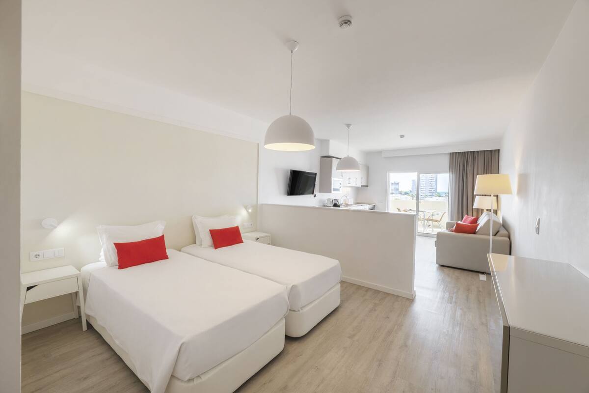 A modern hotel room with two beds, a desk, a chair, a TV, and a view of the city.