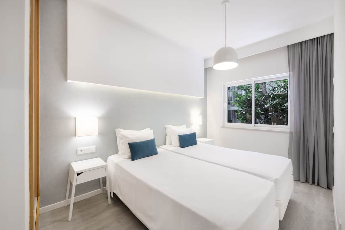 A modern hotel room with two beds, a bedside table, a nightstand, and a large window with curtains.