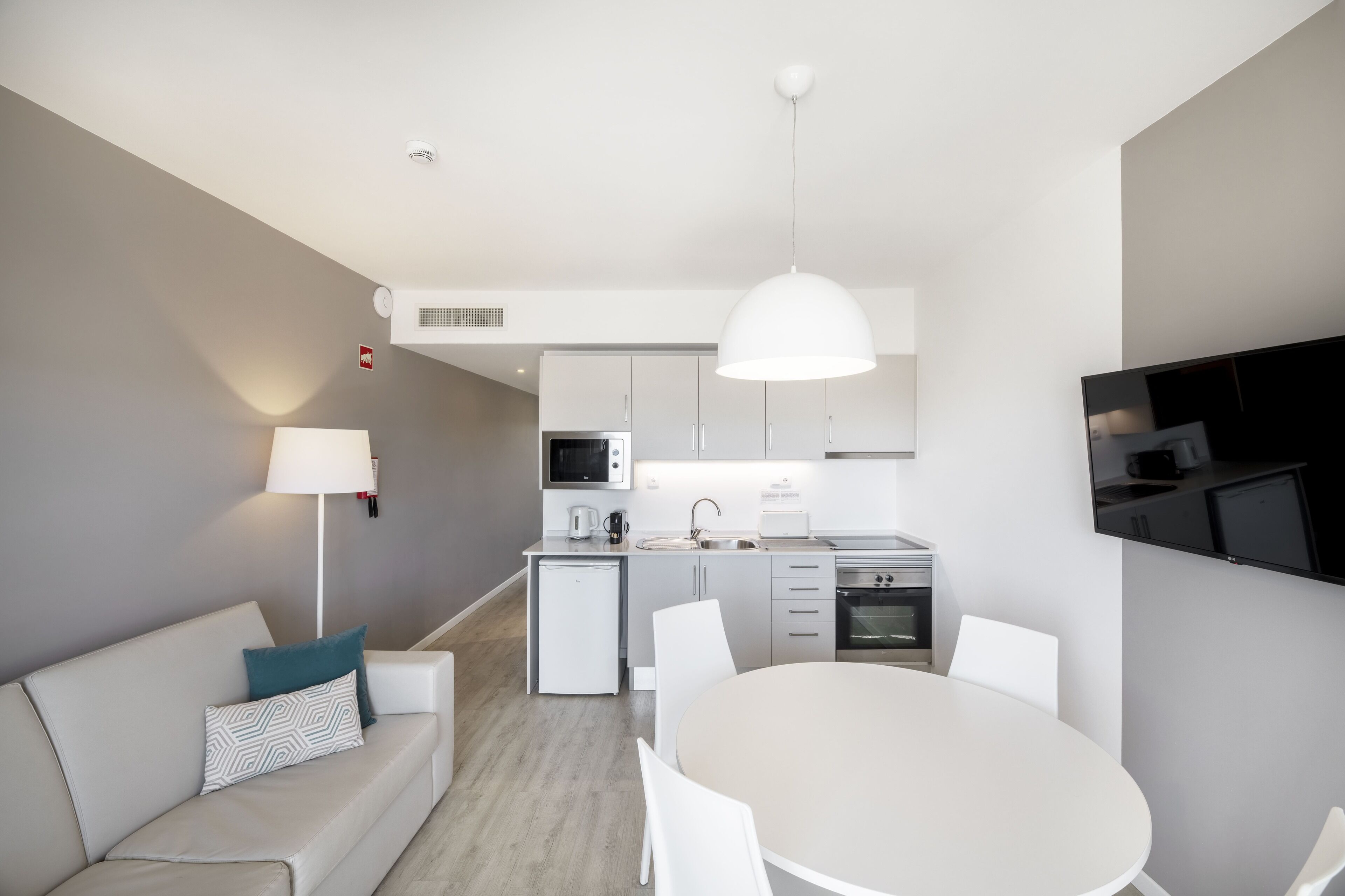 A modern kitchen with white cabinets, a built-in microwave, and a stove. There is a dining area with a round table and chairs, and a sofa with a patterned pillow.