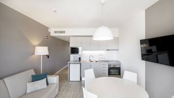 A modern kitchen with white cabinets, a built-in microwave, and a stove. There is a dining area with a round table and chairs, and a sofa with a patterned pillow.