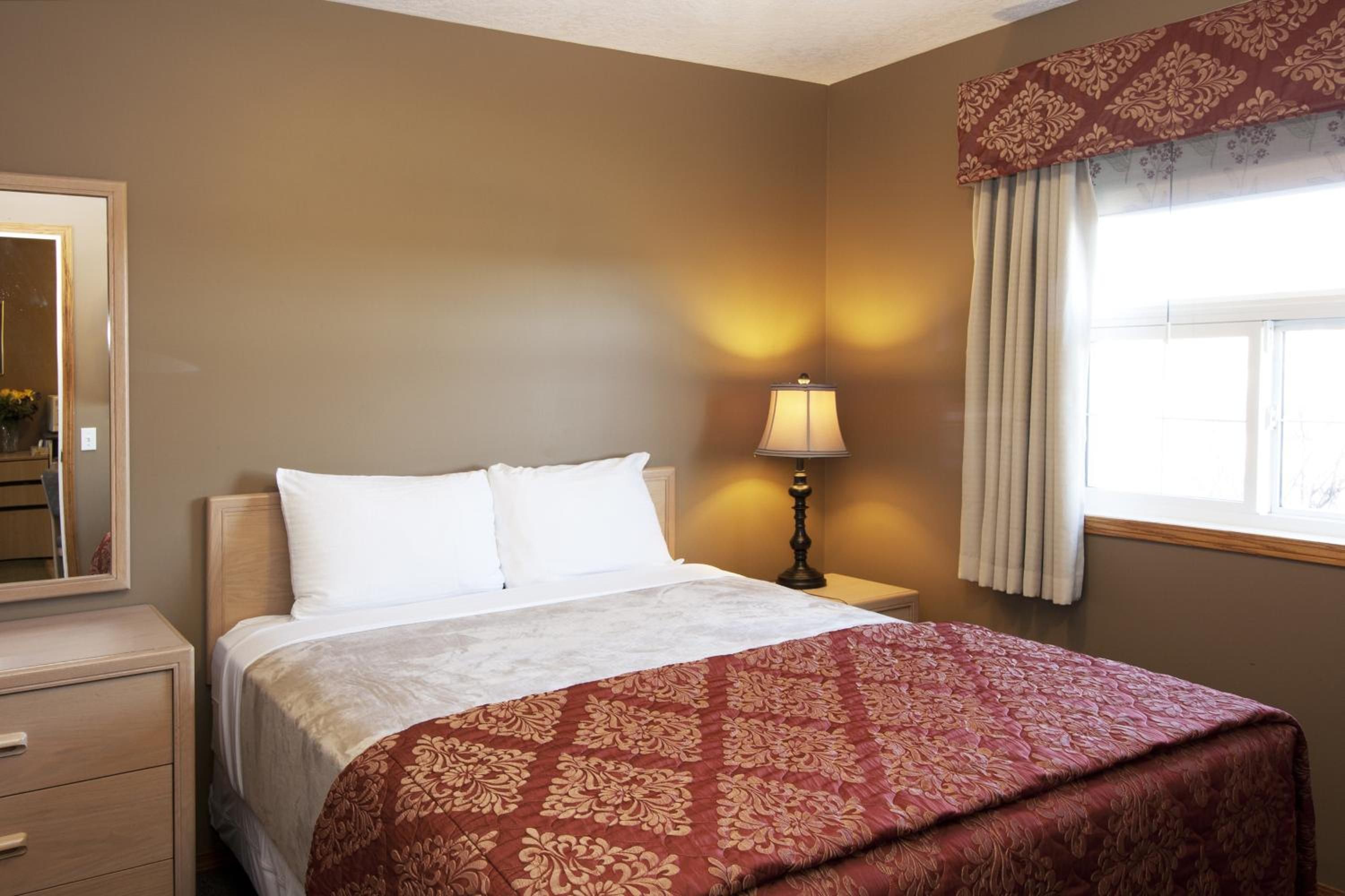 Habitación de hotel con cama, lámpara de noche, cómoda, espejo y ventana con cortinas.