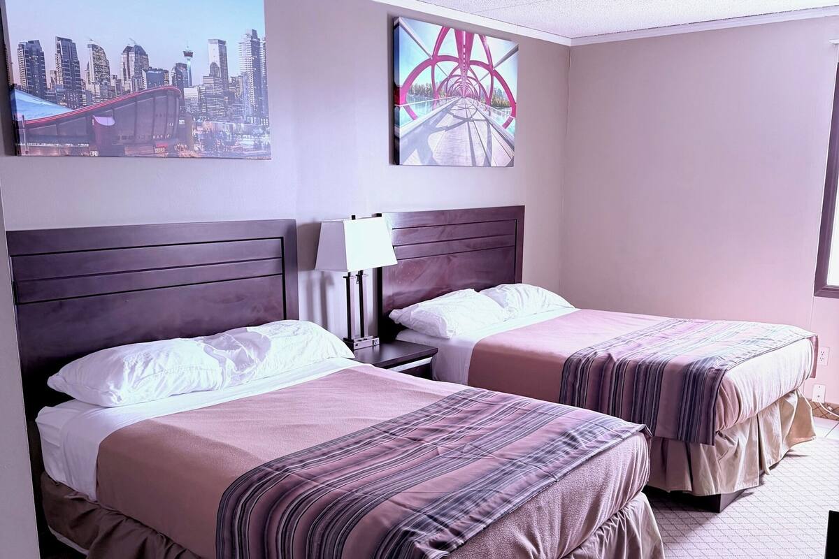 A hotel room with two beds, a cityscape view, a large window, and a cityscape painting.