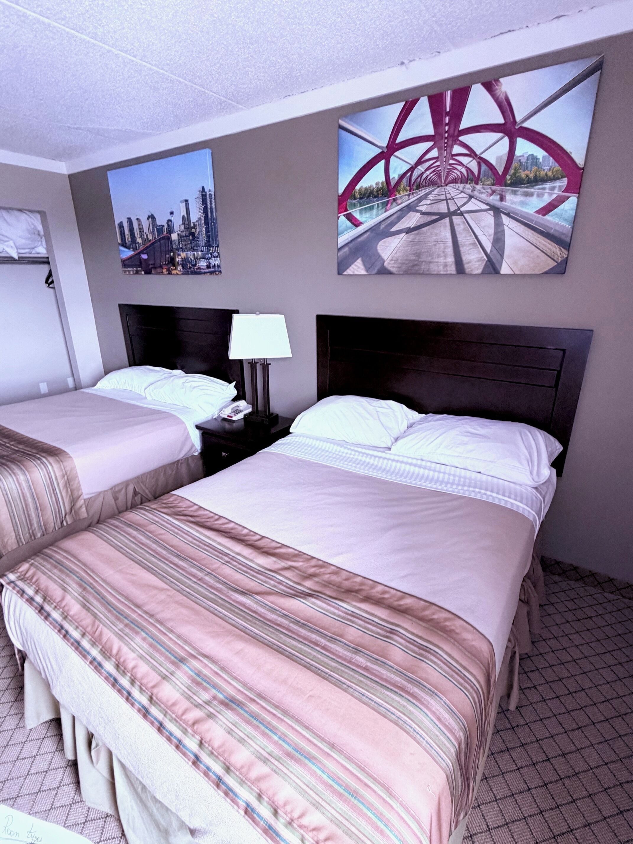 A hotel room with two beds, a cityscape picture, a bridge picture, and a lamp.