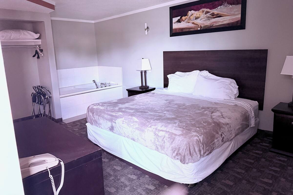 A hotel room with a bed, a nightstand, a lamp, a painting, a bathroom with a bathtub, and a desk with a telephone.