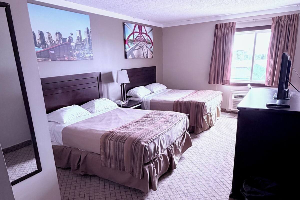 A hotel room with two beds, a desk, a computer, a window with curtains, and a cityscape painting on the wall.