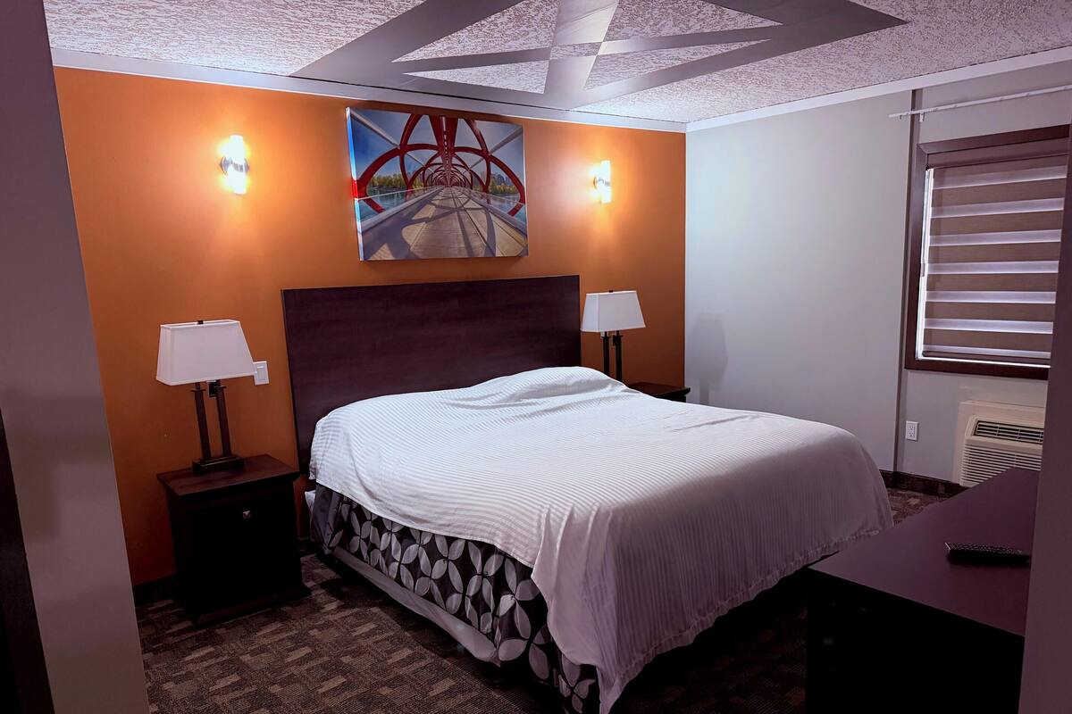 A hotel room with a bed, two bedside lamps, a painting on the wall, and a window with blinds.