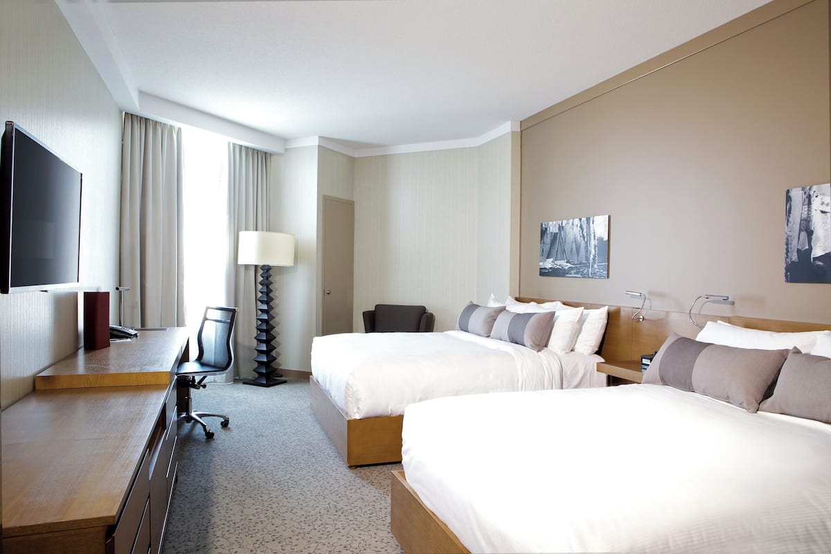 A hotel room with two beds, a desk, a chair, a TV, and a lamp.