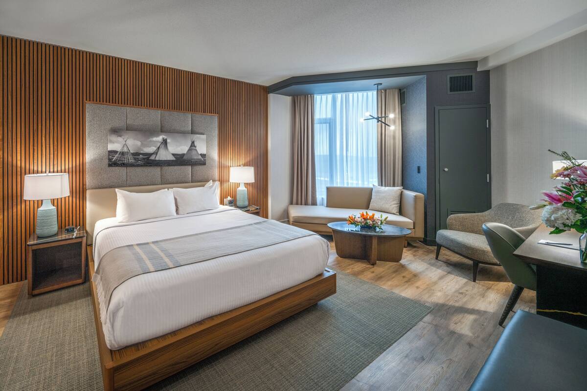 Habitación de hotel con una cama grande, un sofá, un escritorio y una silla. Hay un cuadro en la pared y una ventana con cortinas.