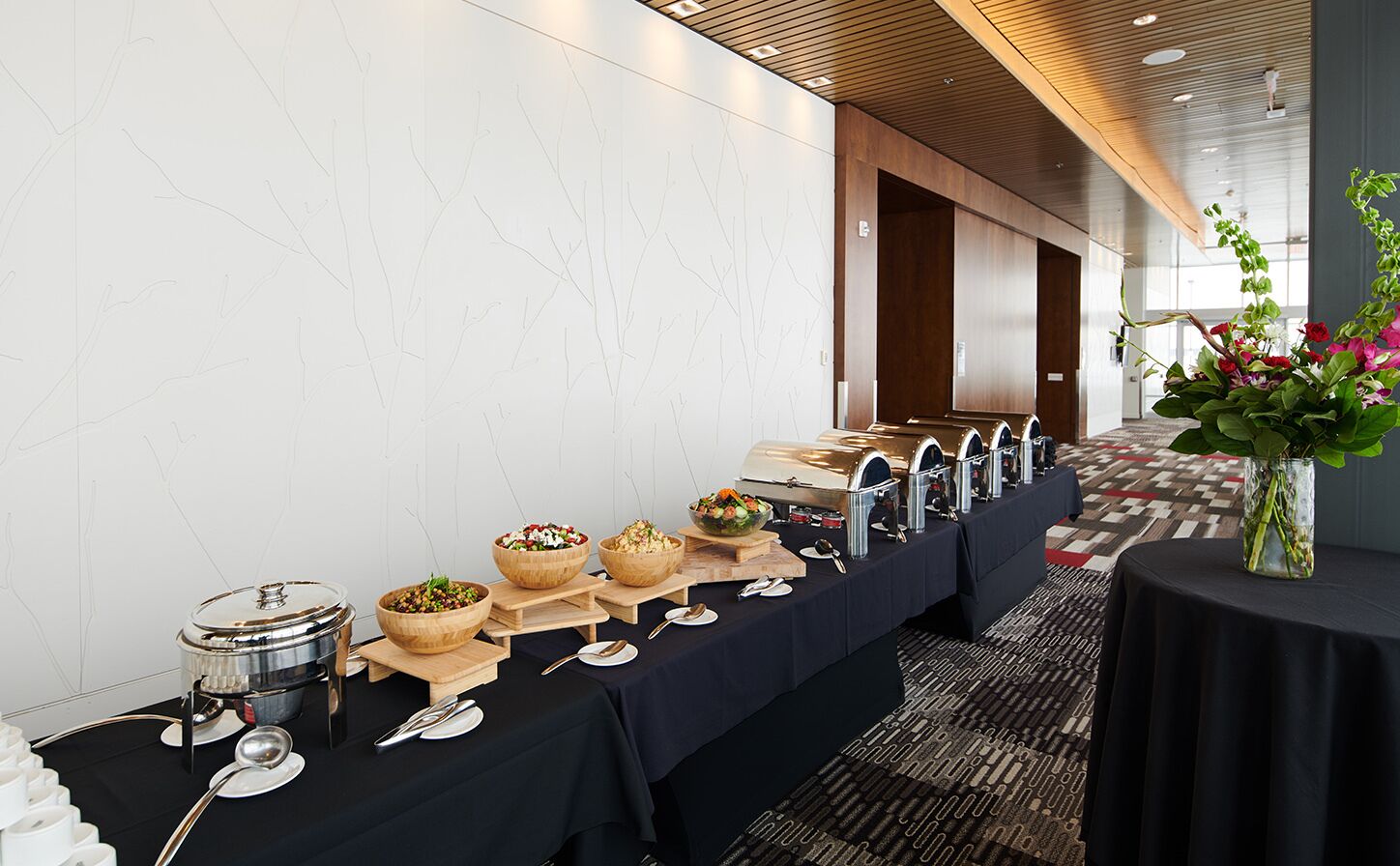 A buffet setup with various food containers and utensils on a long table, set against a modern interior with wooden paneling and a patterned carpet.
