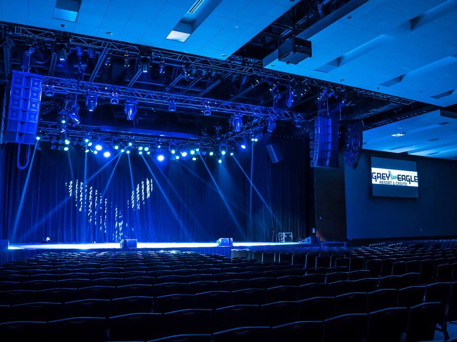 Un auditorio interior con filas de asientos, un escenario con equipo de iluminación y una pantalla mostrando 'Grey Eagle Resort & Casino'.