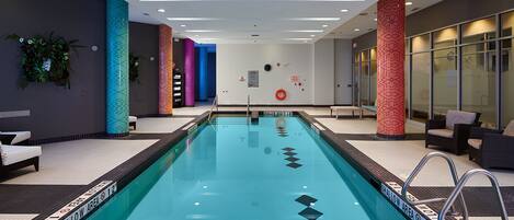 Una piscina interior moderna con línea de agua transparente, rodeada de pilares coloridos y equipada con señales de seguridad y sillones de descanso.