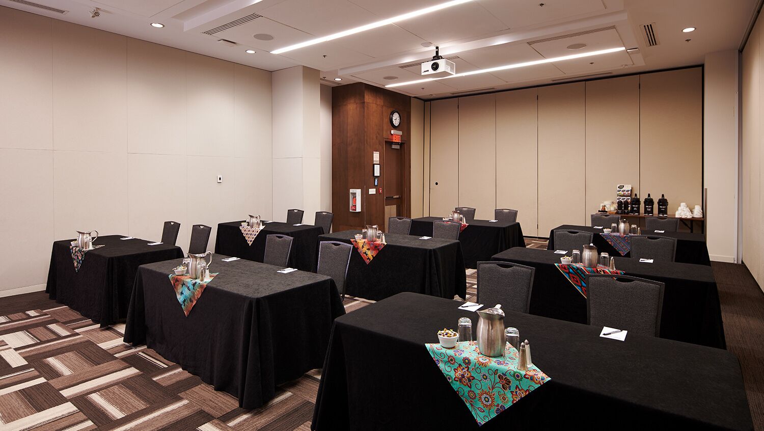 Uma sala de conferências com várias mesas, cada uma com toalha preta e um pequeno objeto decorativo no centro. Cadeiras ao redor das mesas e uma tela de projetor visível no teto.