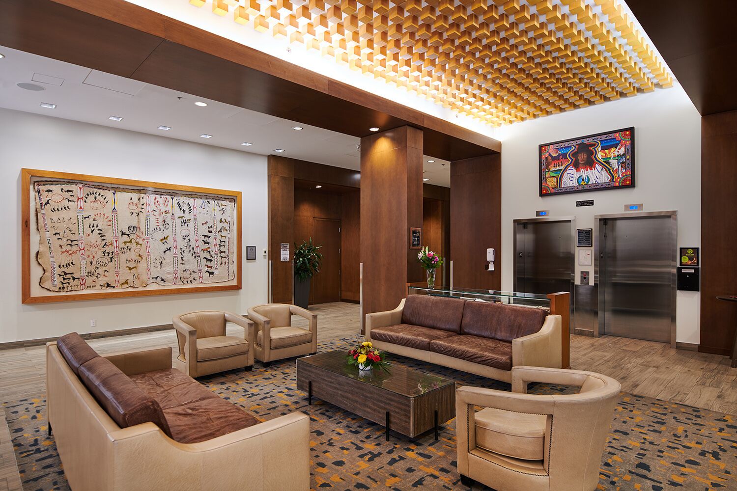 A lobby with a large indigenous artwork, seating area, elevators, and a modern ceiling design.
