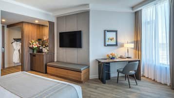 A hotel room with a bed, a desk with a chair, a TV, a framed picture, and a window with curtains.