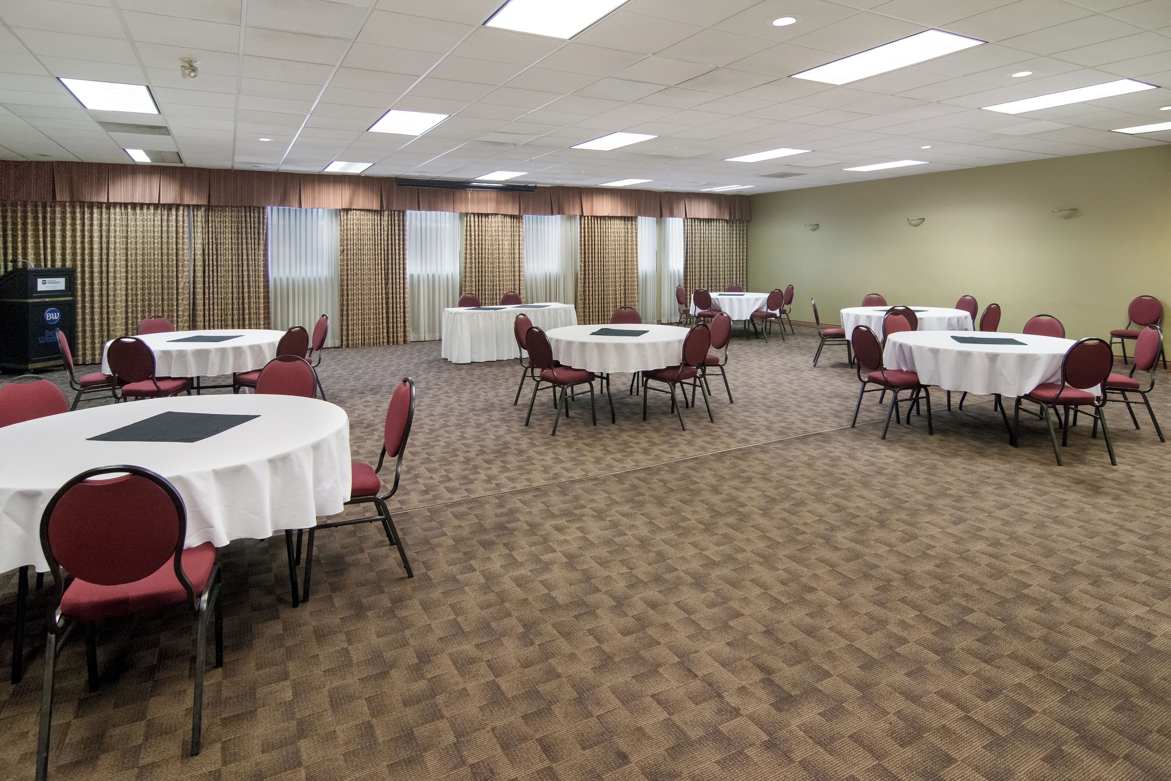 Un salón de conferencias con varias mesas redondas y sillas, piso alfombrado y cortinas en las ventanas.