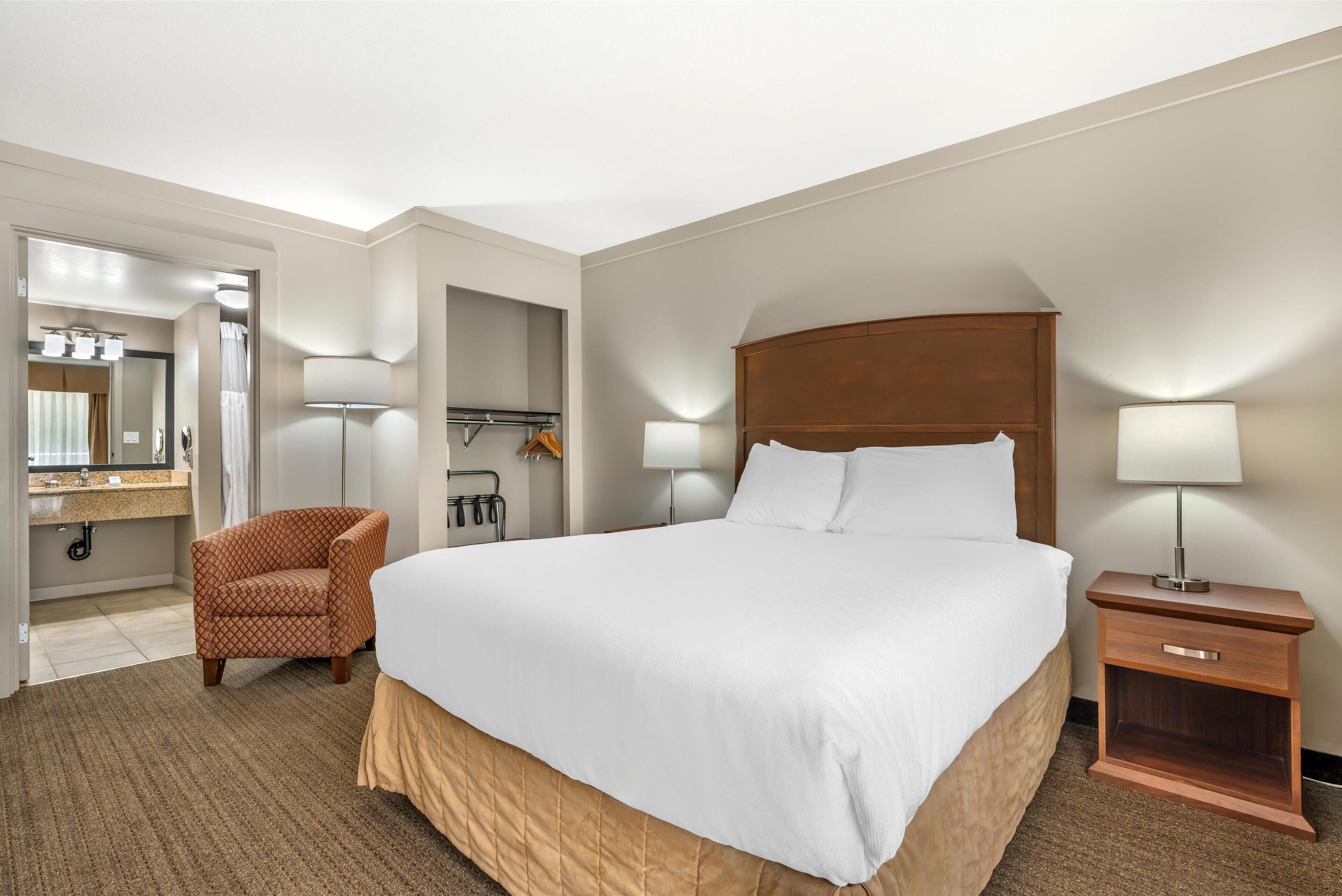 A hotel room with a large bed, a chair, a nightstand, and a bathroom visible through an open door.