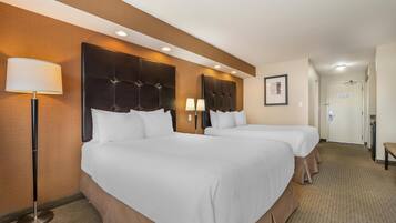 A hotel room with two beds, a large headboard, a floor lamp, and a door in the background.