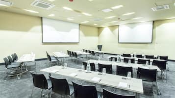 Un salón de conferencias con filas de mesas y sillas, equipado con proyectores y pantallas.
