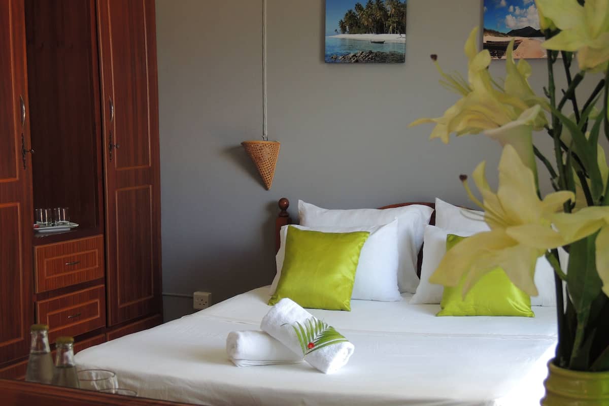 Una cama bien hecha con almohadas blancas y verdes, un armario de madera y una mesita con vasos y botellas.