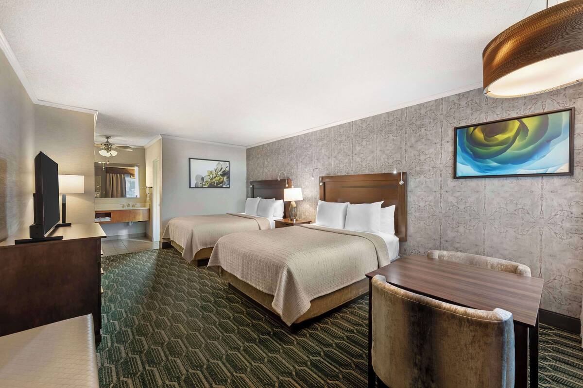 Quarto de hotel com duas camas, uma escrivaninha, uma cadeira, uma televisão, um abajur e um quadro na parede.