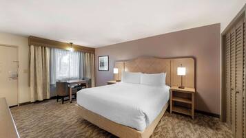 A hotel room with a large bed, a desk, and a chair. There is a lamp on each side of the bed.