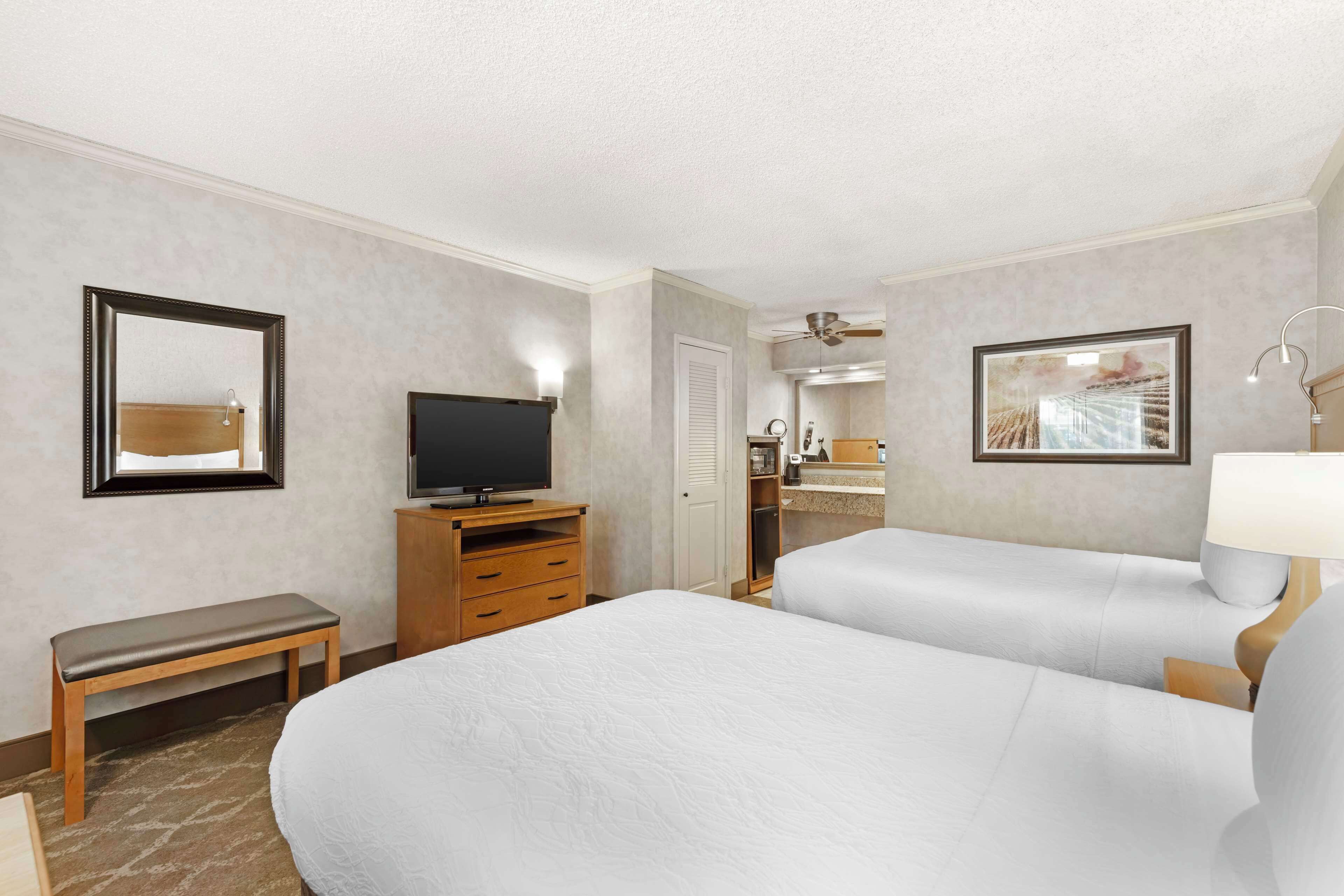 A hotel room with two beds, a TV, a bench, a desk, and a bathroom visible through an open door.