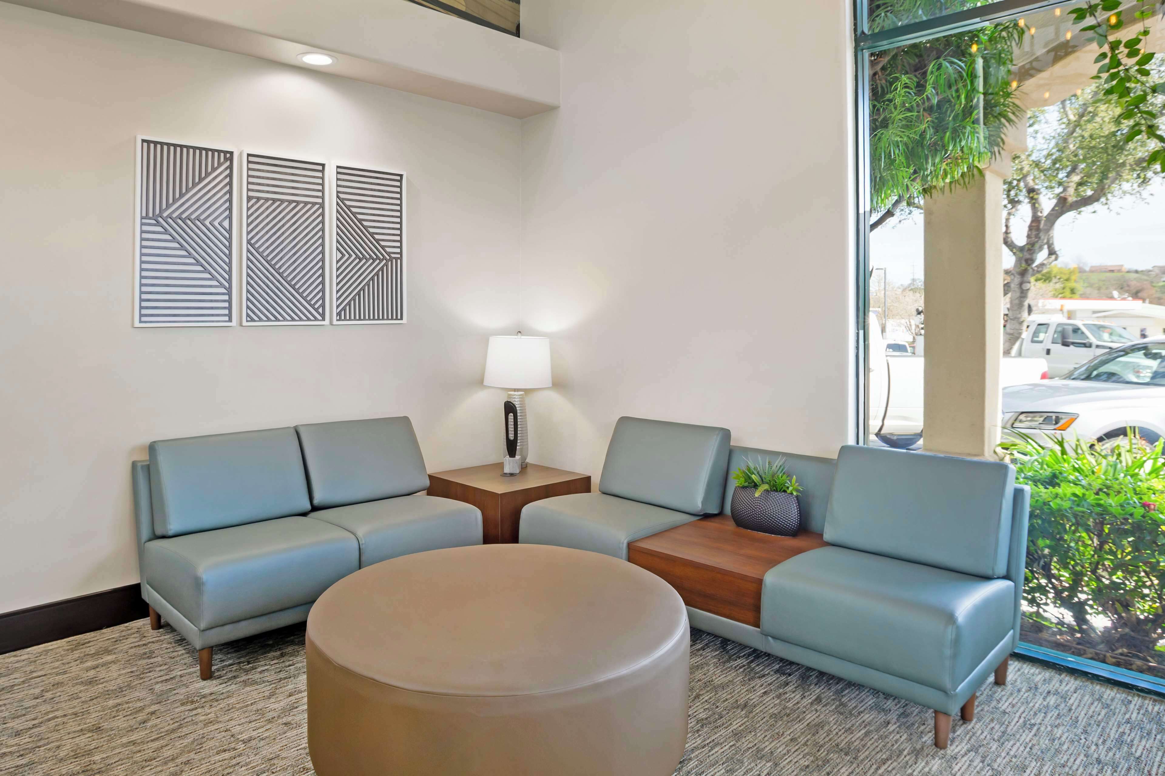 A modern waiting area with a sofa, a round table, and a lamp. There are three wall-mounted geometric art pieces and a window with a view of the outside.