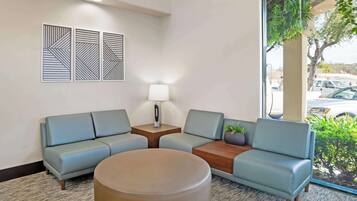 A modern waiting area with a sofa, a round table, and a lamp. There are three wall-mounted geometric art pieces and a window with a view of the outside.