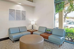 Un espace d’attente moderne, doté d’un canapé, d’une table ronde et d’une lampe. Trois œuvres d’art géométriques sont accrochées aux murs, et une fenêtre offre une vue sur l’extérieur.
