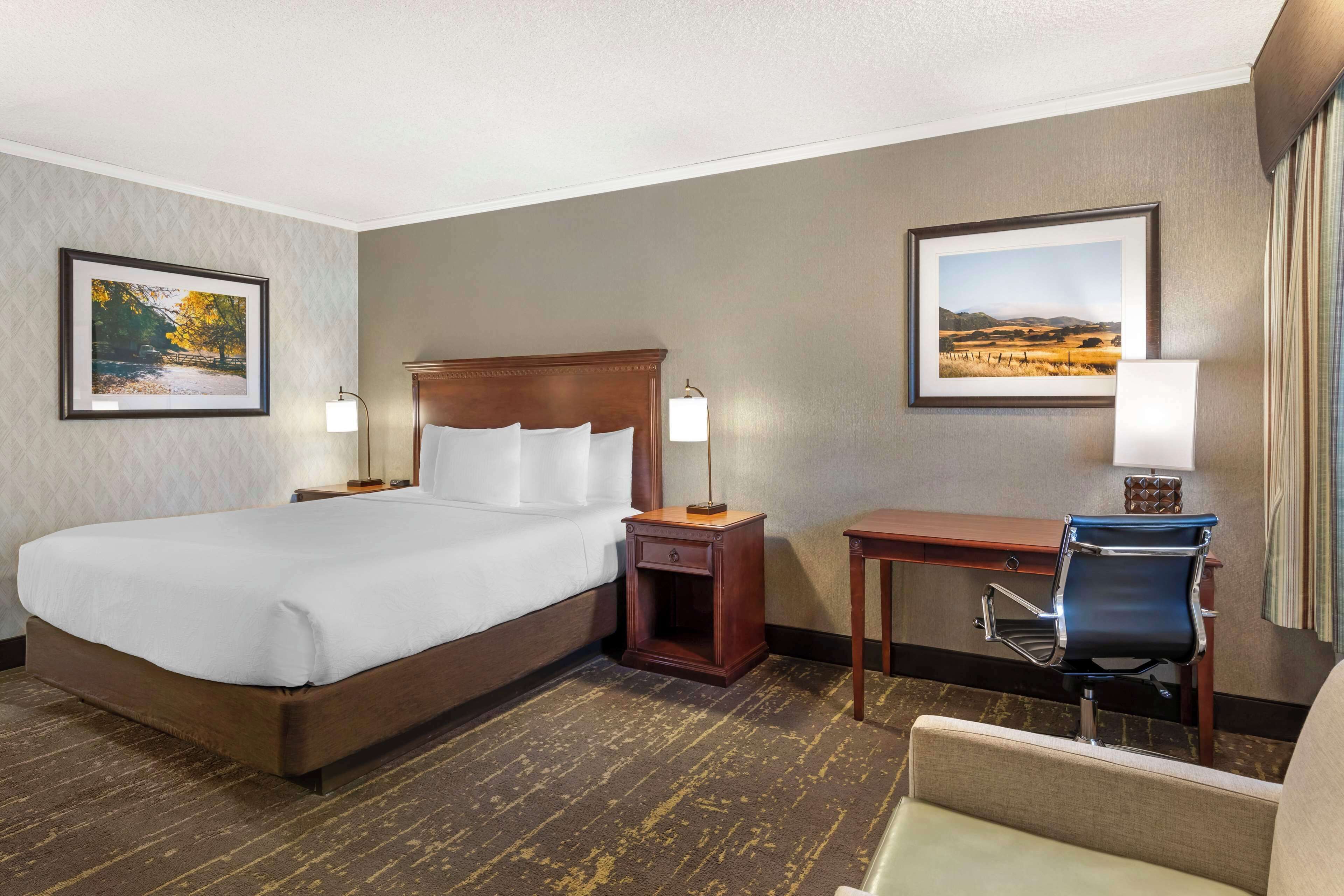 Quarto de hotel com uma cama grande, uma escrivaninha com cadeira, duas luminárias de cabeceira e duas quadros nas paredes.