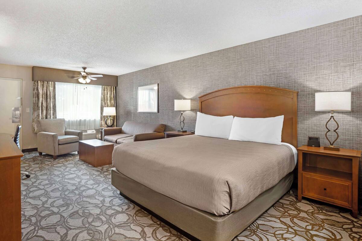 Quarto de hotel com uma cama grande, duas mesas de cabeceira com luminárias, uma cadeira, uma escrivaninha e um ventilador de teto.