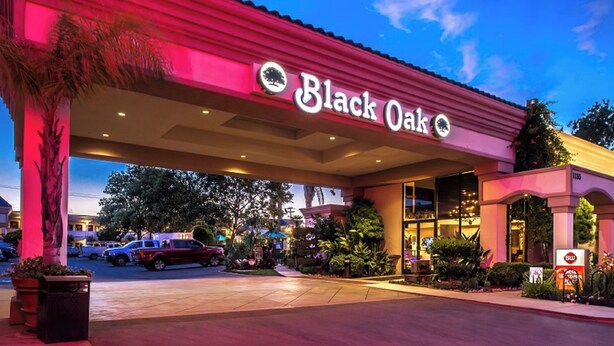 El exterior del hotel Black Oak, con una entrada destacada, un toldo rojo y una zona ajardinada con palmeras y plantas.