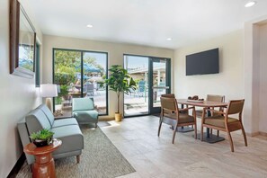 Una sala de estar moderna con un sofá, una mesa de comedor con sillas, un televisor montado en la pared y ventanas amplias con vistas al exterior.