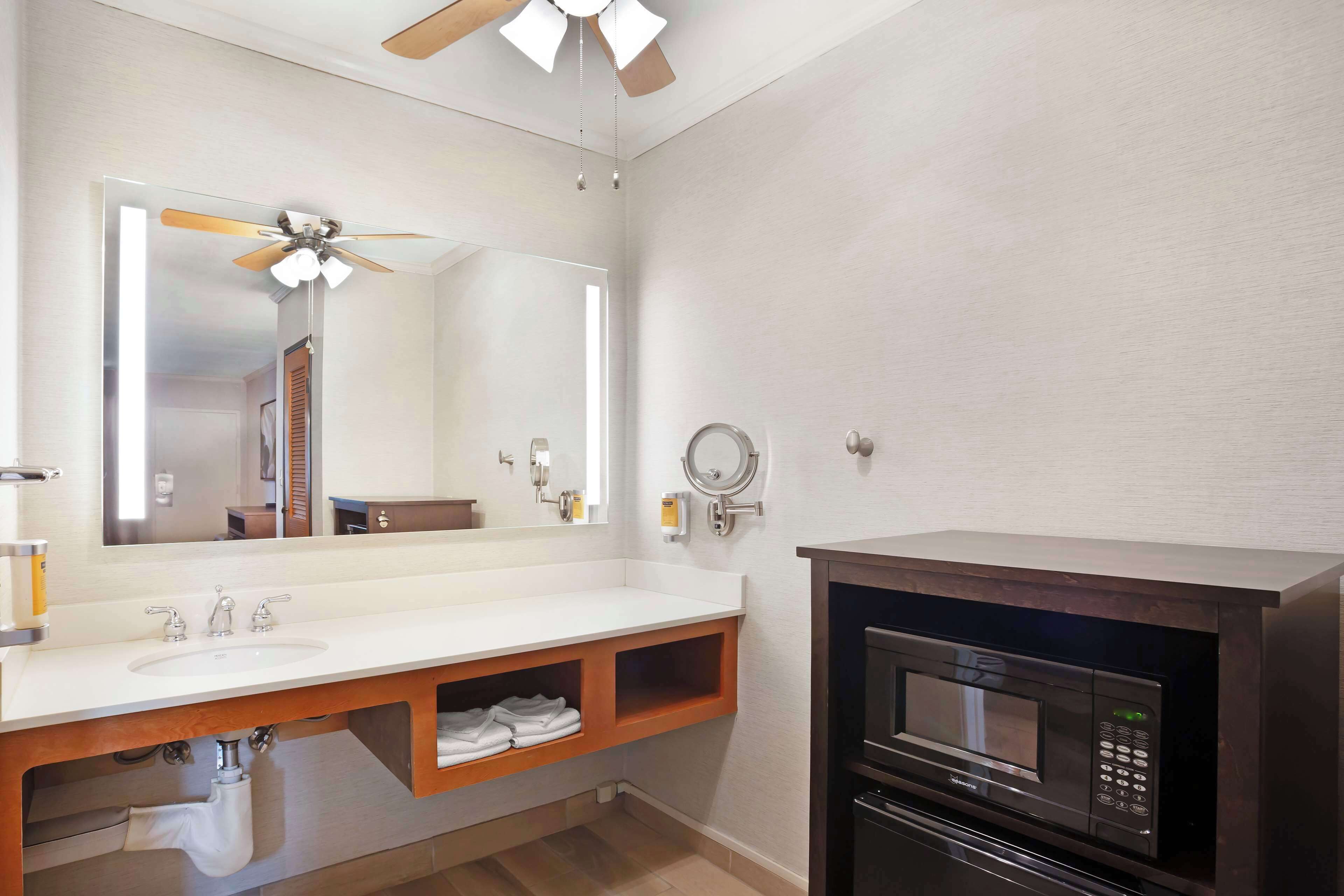 A bathroom with a white sink, wooden vanity, a mirror, a ceiling fan, a microwave, and a towel rack.