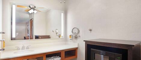 Un baño con lavamanos blanco, tocador de madera, espejo, ventilador de techo, microondas y barra para toallas.