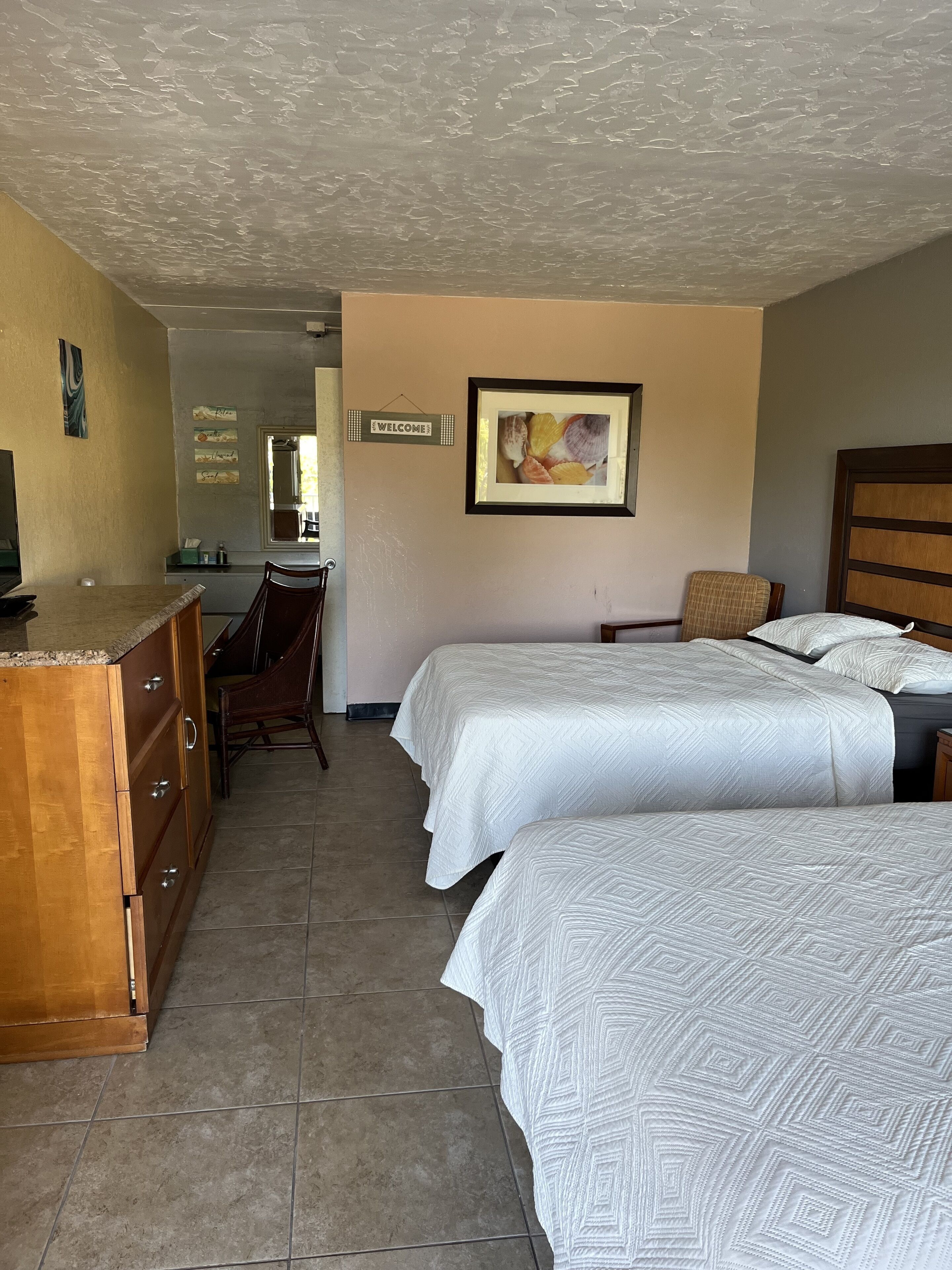 A hotel room with two beds, a wooden dresser, a desk, and a framed welcome sign.