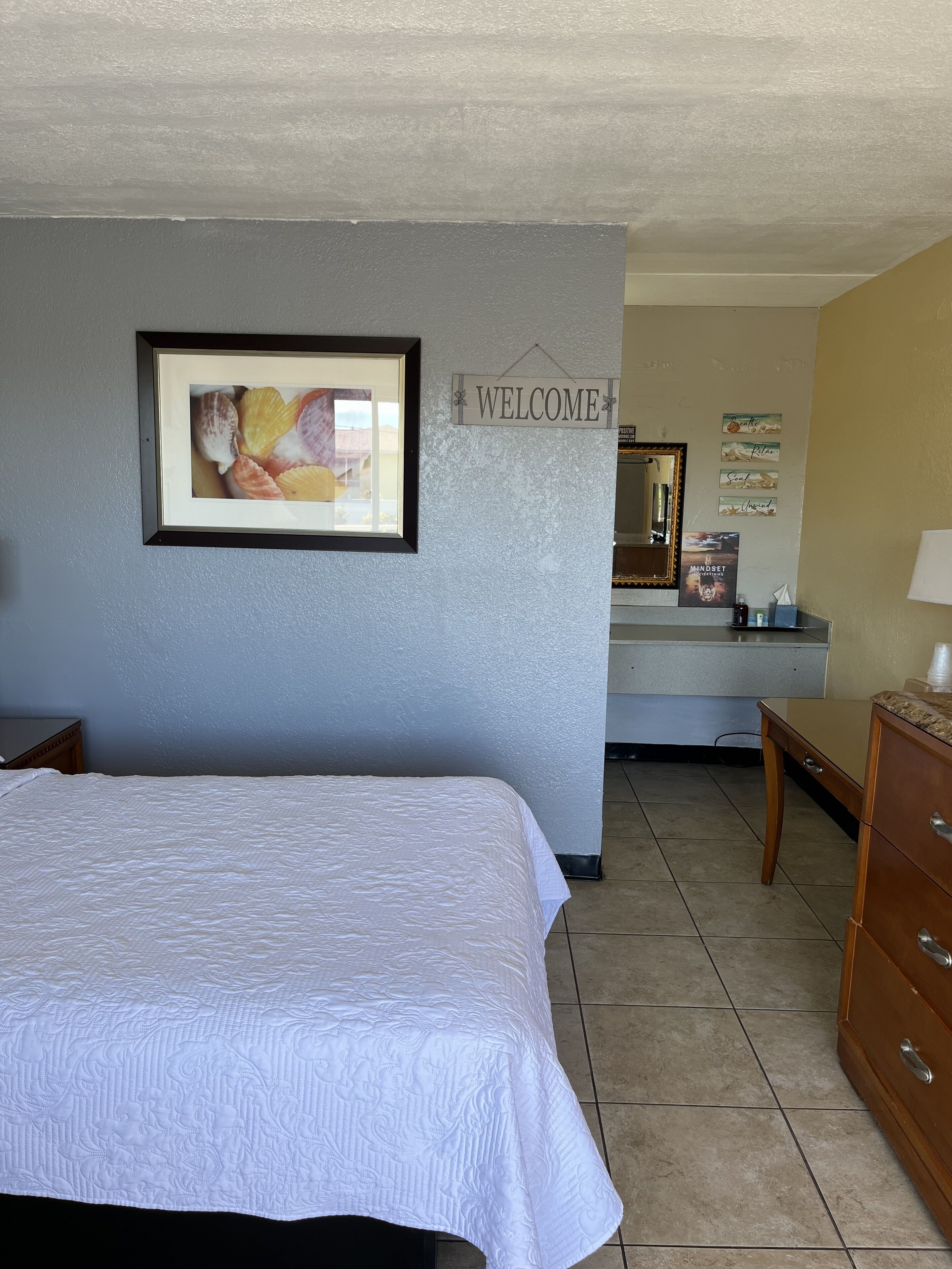 Une chambre d’hôtel comprenant un lit, un bureau, une commode, un panneau d’accueil encadré, un miroir et un mur orné de citations inspirantes.