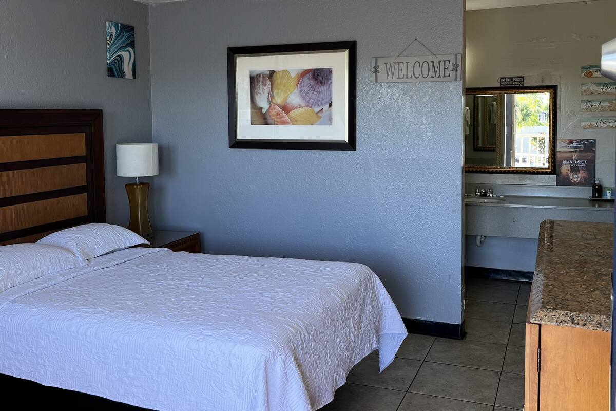 Habitación de hotel con cama, mesita de noche, cuadro enmarco, cartel de bienvenida y vistas al exterior.