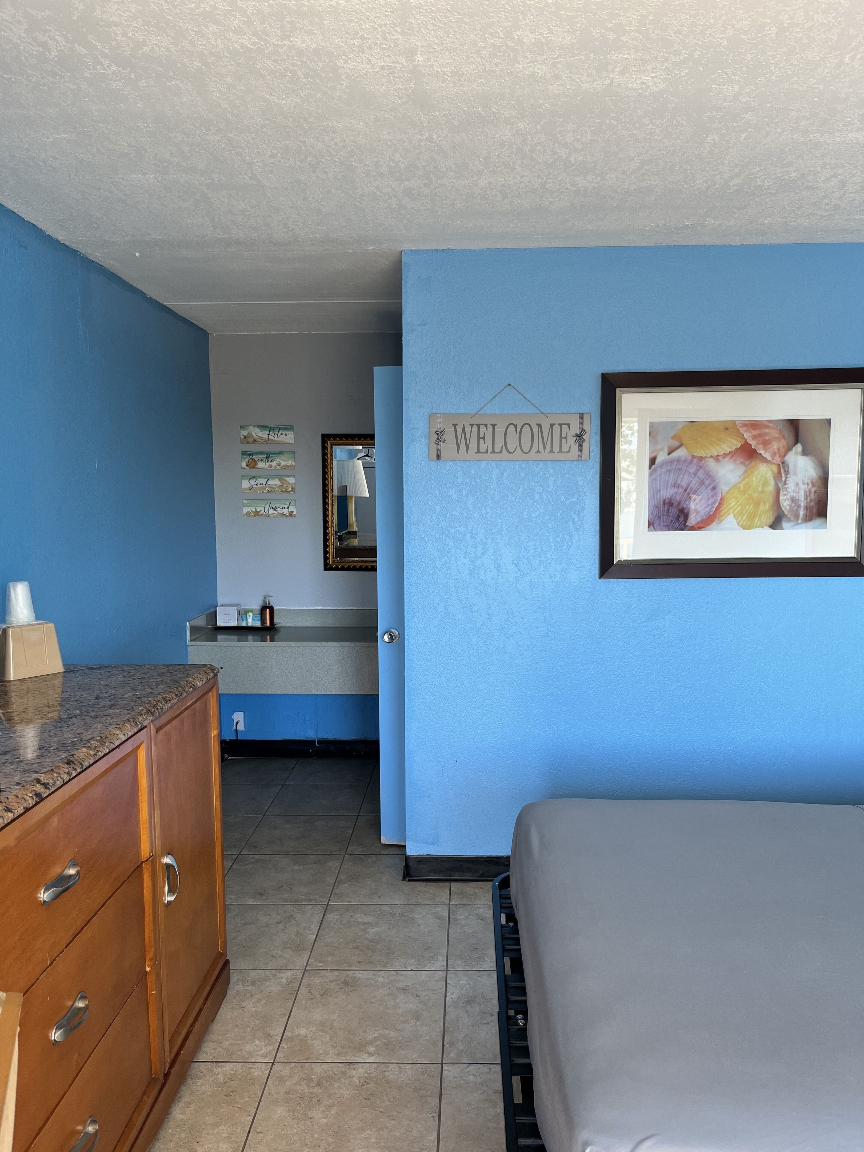 Une chambre d’hôtel avec un mur bleu, un panneau d’accueil, un tableau encadré représentant des coquillages, une commode en bois et un lit.