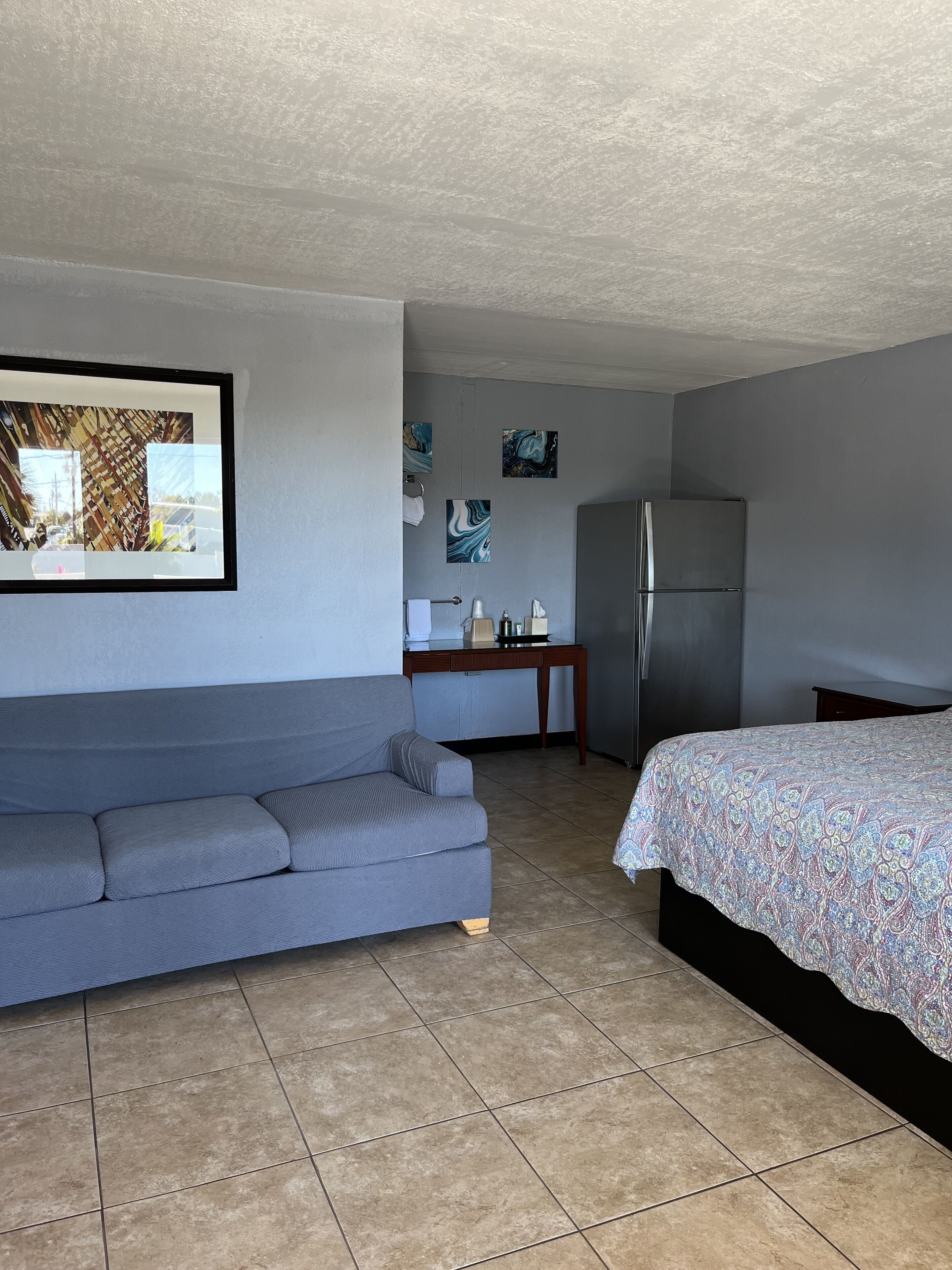 Een compacte hotelkamer met een grijze bank, een bed met een bedovertrek met patroon, een koelkast, een klein tafeltje met toiletartikelen, en een muur met abstracte kunst.