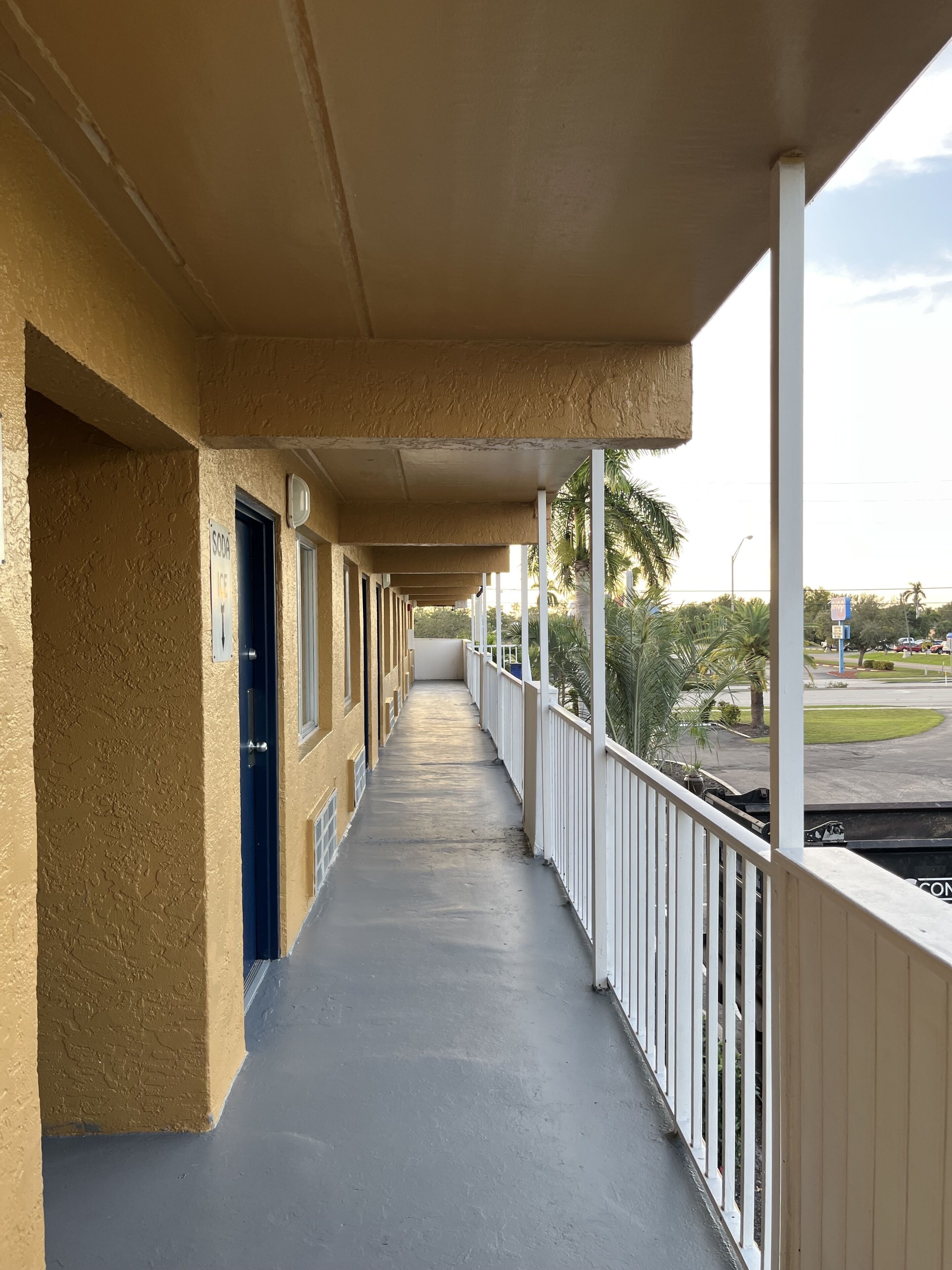 A long corridor with a balcony, white railings, and a view of palm trees and a road.