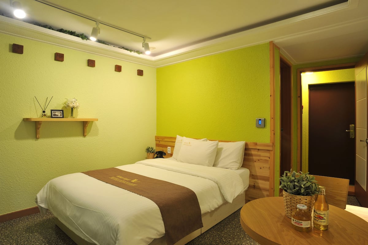Habitació d'hotel amb un llit, una taula de nit amb una ampolla i un bocal, una petita estant amb un quadre i una planta, i un panell de capçal de fusta.