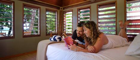 A bedroom with a wooden ceiling, large windows with shutters, a bed with white linens, and a blue and white striped pillow.