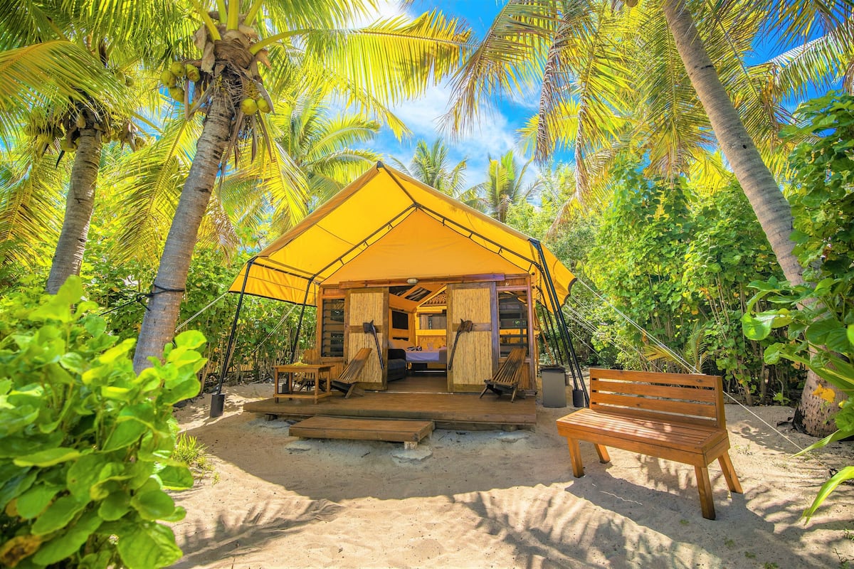 A yellow tent structure with a wooden bench and a table, surrounded by palm trees and sand.